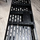 Černý kovový pedalboard s mřížkovým dnem a vyvýšenými okraji. Povrch je matně černý, v pravé části se nachází menší vyvýšená plocha s otvory. Na nást…