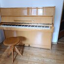 Světle hnědé dřevěné piano s přiloženou kulatou taburetkou. Nad klávesnicí je v dřevě vyryt nápis SCHOLZE. Povrch nástroje vykazuje mírné odrazy svět…