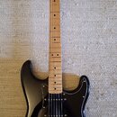 Černá elektrická kytara s jasným lakem a hnědým hnedým hmatníkem. Na hlavě je nápis Squier STRATOCASTER. Kytara má tři snímače, tři ovládací knoflíky…