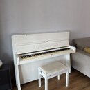 Bílé piano s doprovodnou bílou stoličkou s béžovým polstřením. Na přihradce nad klávesami je vidět nápis PETROF. Nástroj vykazuje mírné opotřebení v…