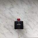 Černý hranatý adaptér s červeným knoflíkem v horní části. Na přední straně jsou nápisy USB a DC 12V 300mA následovaný symbolem polarity. V dolní část…