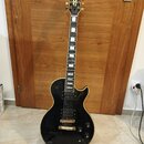 Černá elektrická kytara s gold hardwarem, umístěná na černém stojanu. Na hlavě je zlatý nápis Gibson. Nástroj má dva humbuckery, dva ovládací knofíky…