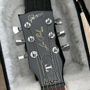 Černá hlava elektrické kytary v otevřeném kufru s bílou výplní. Na hlavě je bílý nápis Gibson a pod ním model Les Paul MODEL. Ve spodní části hlavy j…