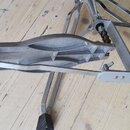 Fotografie zobrazuje kovový mechanismus basového pedálu v stříbrné barvě. Pedál má plastovou stupnici šedé barvy a je vybaven kovovými tyčemi s černý…