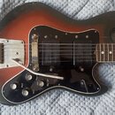 Elektrická kytara s tělem v červeno-černé barvě (sunburst) a černým pickguardem. Nástroj má dva humbuckery, kovový most s páčkou a čtyři ovládací kno…