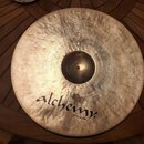 Kovový činel zlatavé barvy s viditelným rakováním povrchu. Na spodní části je černý nápis alchemy cymbals®. V horní části jsou drobné, lehce čitelné…