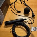 Černý hlavový mikrofon s čelníkem a černou molitanovou windscreen. Na přívodním kabelu je umístěn černý adaptér s bílým nápisem MPA VL MICRO MIC PHAN…