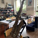 Stříbrný saxofon umístěný na černém stojanu značky HERCULES. Nástroj vykazuje výrazné opotřebení povrchu, oxidaci a zahlreiche škrábance. Na těle nás…