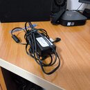 Černý síťový adaptér s napájecím kabelem a konektorem. Na boku adaptéru je bílá nálepka s technickými specifikacemi, z nichž jsou čitelné nápisy AC I…