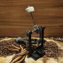 Černý pedál basového bubnu s kovovými mechanismy a řetězem. Na horní části je připevněna kovová tyč s bílým molanovým tlumicím blokem. Na nástroji ne…