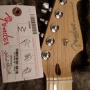 Fotografie zachycuje hlavu elektrokitary z jasného dřeva s chromovanými ladičkami. Na hlavě je černé logo Fender CORONA CA a nápis STRATOCASTER®. Ved…