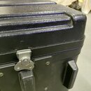 Detail černého tvrdého kufru s texturovaným povrchem. Na přední straně je vidět kovový stříbrný zámek a dvě černé plastové rukojeti. Povrch vykazuje…