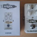 Bílá efekt pedál s nápisy SPARK BOOSTER a tc electronic. Pedál má čtyři bílé ovládací knoflíky s popisky GAIN, LEVEL, BASS a TREBLE, dále přepínač FA…