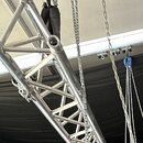Detail stříbrného hliníkového systémového trussu s mřížkovou konstrukcí. Vedle konstrukce jsou vidět kovové řetězy a modrý zvedací mechanismus. Na po…