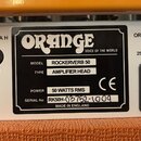 Detail zadní strany kytarového komba s bílým panelem a oranžovým povrchem. Uprostřed je černý štítek s bílým logem ORANGE a nápisem VOICE OF THE WORL…