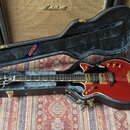 Červená elektrická kytara v černém tvrdém kufru s modrým nápisem GRETSCH. Na hlavě kytary je bílé logo GRETSCH a na hmatníku jsou viditelné vložky. K…