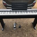 Černý digitální piano s klaviaturou a přiloženým notovým pultem na černém stojanu. Na pravé straně horního panelu je nápis CASIO PRIVIA. Pod nástroje…
