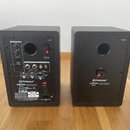 Dva černé studiové monitory v kostkovém tvaru s nápisy Left a Right. Na zadní straně levého monitoru jsou ovládací prvky Acoustic Tuning, přepínač Ou…