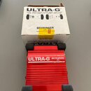 Červená DI box s hliníkovým žebrovaným povrchem a černými gumovými nohami. Na zařízení je bílý nápis ULTRA-G, logo BEHRINGER a text ACTIVE DI-BOX/VIR…