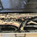 Stříbrný saxofon v černém polstrovaném kufru. Na korbide je viditelný nápis "S.Y.A.N. PARIS". Vedle nástroje je umístěn munční dutý hnedý mouthpiece…