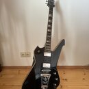 Černá elektrická kytara s atypickým, ostře hranatým tvarem korpusu a hmatníkem s ozdobnými vklády. Na hlavě je viditelné logo "Schecter". Nástroj má…