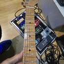 Elektrická kytara s přírodním dřevěným povrchem hředníku a hlavy. Na hlavě je viditelné logo Squier. Kytara je držena v ruce nad efektovou pedalboard…