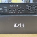 Černý audio interface značky AUDIENT položený na černé krabici s nápisem iD14 10in | 6out Audio Interface. Zadní strana zařízení obsahuje USB-C port,…
