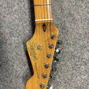 Detail hlavy dřevěné kytary s nápisy Squier, BY FENDER a STRATOCASTER®. Na hlavě jsou umístěny šest chromovaných ladiček. Povrch dřeva je v přirozené…