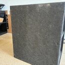 Fotografie zachycuje zadní stranu černého reproduktorového boxu potaženého texturovaným kobercovým materiálem. Povrch vykazuje známky opotřebení v po…