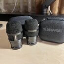 Dvě černá dynamická mikrofony s mřížkou a kovovým tělem. Na obou je viditelný nápis SHURE, na pravém mikrofonu je dále drobný nečitelný nápis začínaj…