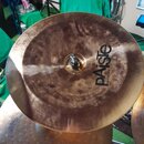 Kruhová činely v bronzové barvě s viditelnými stopy opotřebení a nečistotami na povrchu. Na činely je černý nápis pAiSte a pod ním menší text MADE IN…