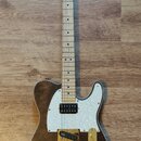 Elektrická kytara typu Telecaster s hnědým dřevěným povrchem těla a bílým perleťovým pickguardem. Nástroj má zlatý hardware, včetně ladíček a mostu,…