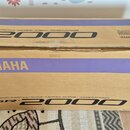 Kartonová krabice s modrým pruhem, ve které je elektronický klavír. Na krabici jsou viditelné nápisy YAMAHA a PSR-2000, doprovázené textem ELECTRONIC…