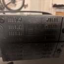 Černý obdélníkový přístroj z plastu s ventilačními mřížkami na bocích. Uprostřed je nápis BATTERY a text DC 12V 1.5Y X 8P SIZE A LFF, DC 9V 12Y X 6P…