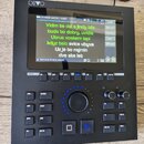 Černý elektronický přístroj s displejem a ovládacími tlačítky. V horním levém rohu je bílý nápis DIVO. Na displeji je zobrazen text písní s nadpisem…