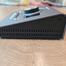 Šedý elektronický přístroj s kovovým povrchem a hranatým tvarem, který se směrem k zadní části svažuje. Na horní straně je umístěn velký kulatý ovlád…