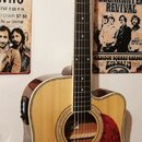 Akustická kytara s přístupem pro praváky, přírodní barva těla a tmavě hnědá hlava. Na hlavě je viditelné logo Epiphone. Kytara má červený pickguard a…