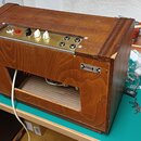 Dřevěný rádiový nebo zesilovačový přístroj s lakovaným povrchem. Na horním ovládacím panelu jsou umístěny dva bílé knofíky, jeden černý knofík, červe…