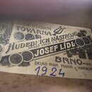 Fotografie detailu starého papírového štítku s nápisy. Je na něm uveden text: „C. AK DVORNÍ TOVÁRNA HUDEBNÍCH NÁSTROJŮ PRISEZ. ZNALEC C. K. ZEM. SOUD…