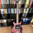 Čtyřstrunná basová kytara s červeným trupem a hnědým hmatníkem. Na hlavě je zlaté logo Gibson a nápis Gibson Custom Shop. Instrument má černý pickgua…