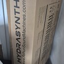 Hnědý papírový karton s nápisem HYDRASYNTH a webovou adresou www.AshunSoundMachines.com. V dolní části je logo ASM s textem ASHUN SOUND MACHINES. Na…
