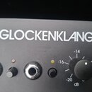 Detailní pohled na černý panel hudebního zařízení s bílým nápisem GLOCKENKLANG. Jsou vidět dva ovládací knoflíky, dvě zásuvky a dvě kontrolní LED dio…
