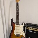 Elektrická kytara s tónem sunburst, bílým pickguardem a javorovou hlavou. Na hlavě je viditelné logo Fender. Vedle kytary stojí černý kytarový zesilo…