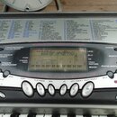 Detailní pohled na ovládací panel digitálního klavíru. V horní části je umístěn seznam skladeb s nadpisem SO SONG BANK a dalšími kategoriemi. Uprostř…