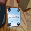 Detail spoje krku a těla kytary z dřeva s viditnou masivní strukturou. Na spoji je přišroubovaná kovová plácačka s nápisy MATSUMOKU a MADE IN JAPAN.…