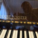 Detail klaviatury a horní části pianina z tmavého lesklého dřeva. Na povrchu je bílý nápis AUGUST FORSTER a pod ním menší text CZECHOSLOVAKIA. Pod ná…