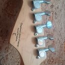 Fotografie zachicuje hlavu kytary z přírodního světlého dřeva s šesti chromovanými ladičkami. Na zadní straně hlavy je černý nápis Tangewood Guitar C…