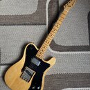 Elektrická kytara s přírodním dřevěným povrchem těla a hnedým javorovým hmatníkem. Na hlavě je viditelné logo "Fender". Černý pickguard obsahuje dva…