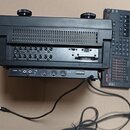 Černý elektronický přístroj s dvěma ovládacími knoflíky nahoře a ventilační mřížkou. Na zadním panelu jsou vstupy a výstupy s nápisy INPUT (čísla 1–8…