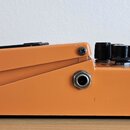 Kytarový efekt pedál v oranžovém provedení. Na boku je vidět jeden vstupní/výstupní konektor a nahoře tři černé oválné kontrolní knoflíky. Povrch je…